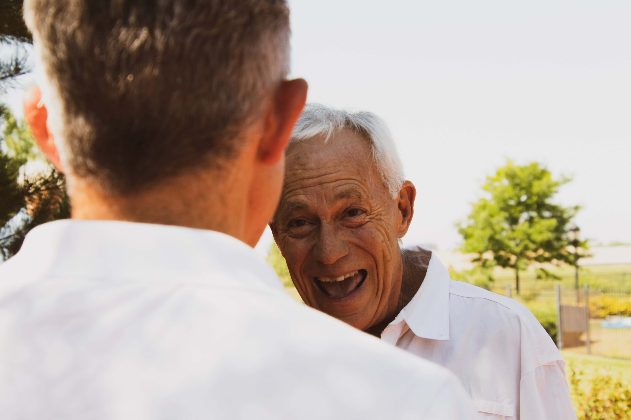 Two elderly men wearing white shirts are outdoors; one facing the camera and smiling, while the other, whose back is to the camera, engages in conversation. Trees and a field are blurry in the background.