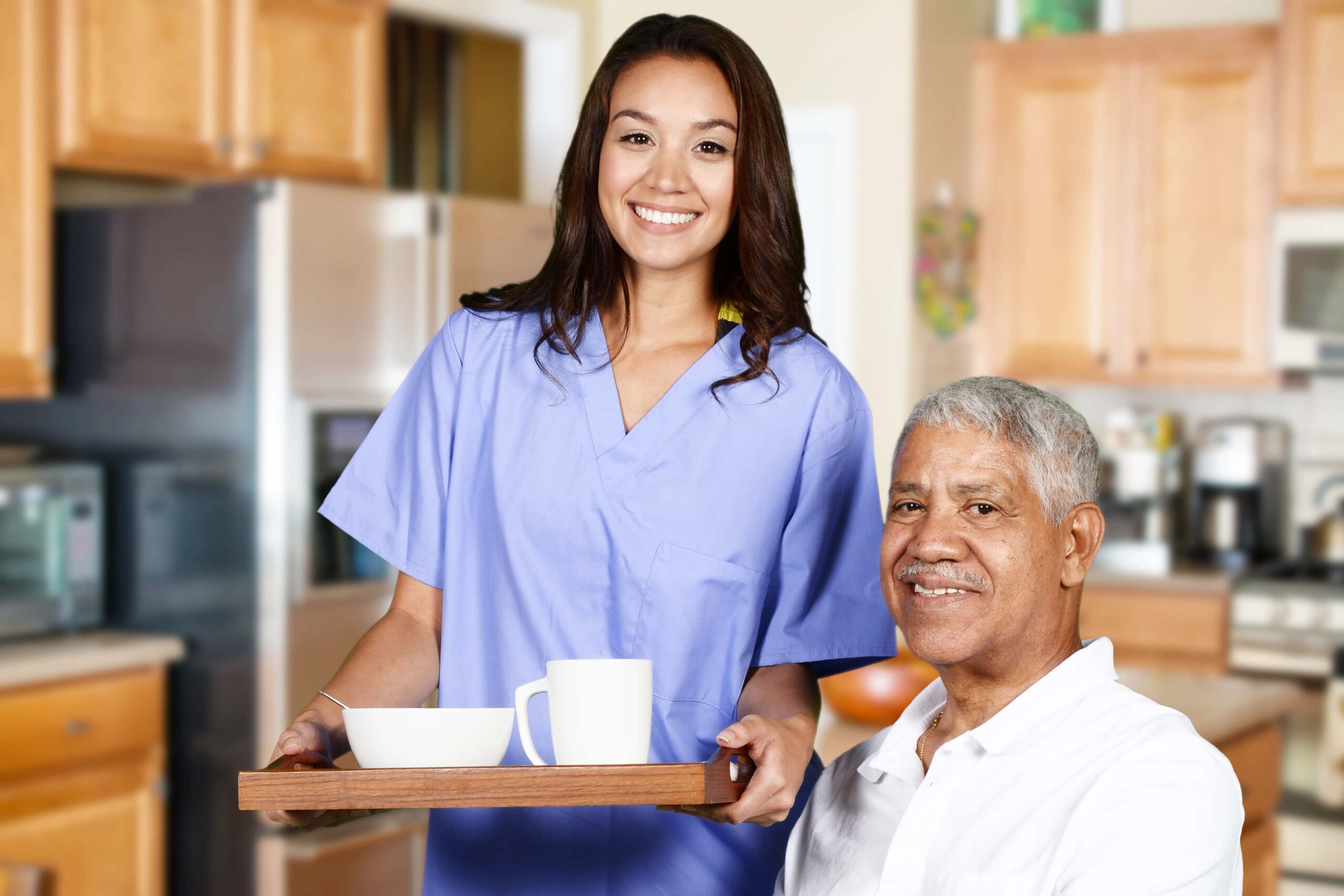 A smiling woman in scrubs holds a tray with a bowl and mug. An older man sits and smiles beside her in a kitchen setting.