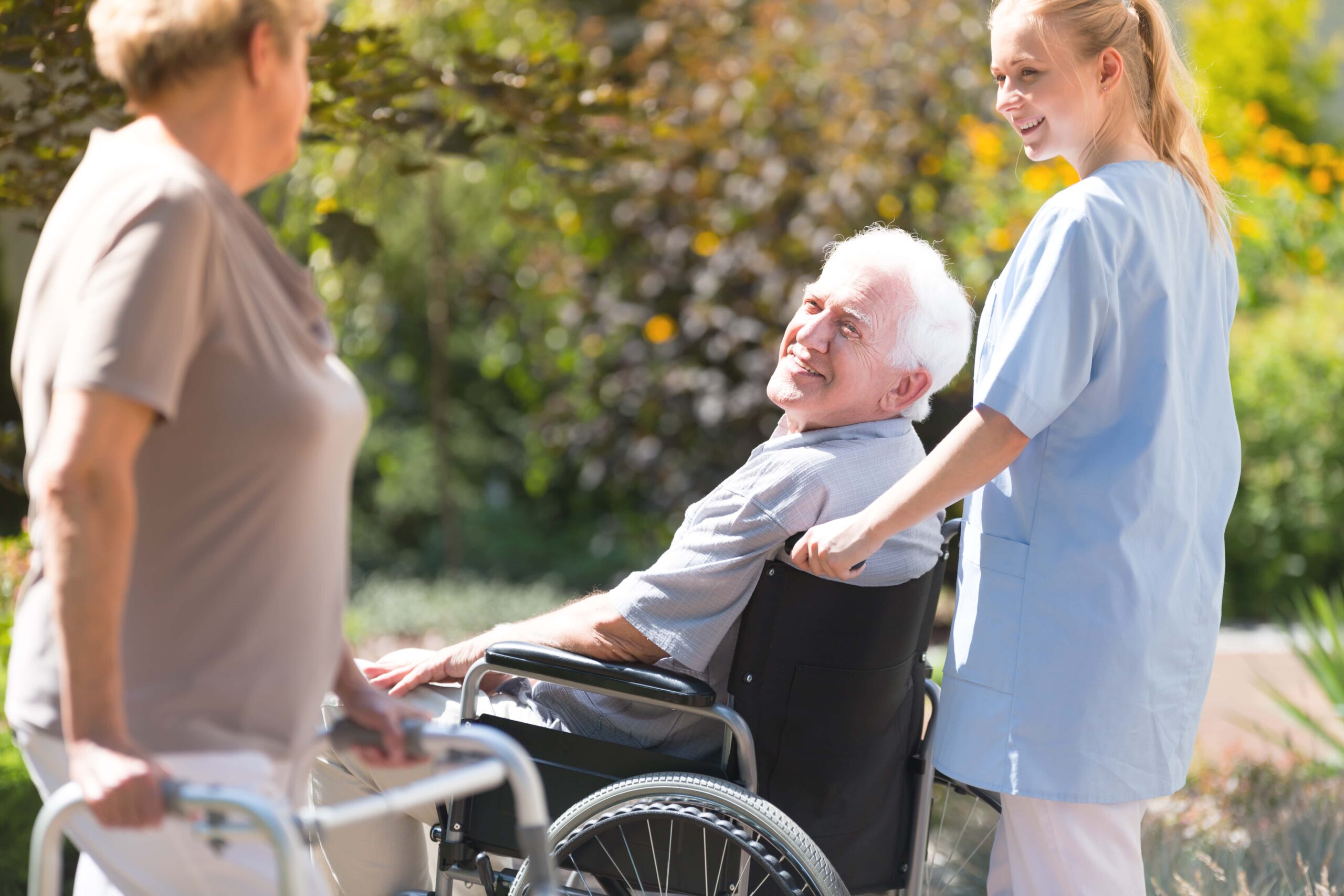An elderly man in a wheelchair is being assisted by a nurse in blue scrubs. Another older person with a walker is in the foreground. They are outdoors in a garden setting.