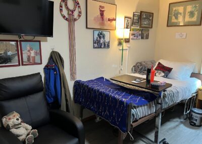A cozy room with a bed, blue decorative cover, wall-mounted TV, framed pictures, black armchair with a teddy bear, and a coat on a chair. Lamp and bedside table present.