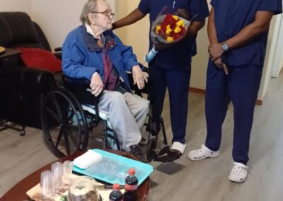 An elderly person in a wheelchair receives a bouquet from a healthcare worker, while another healthcare worker stands beside them. A table with food and drinks is in the foreground.