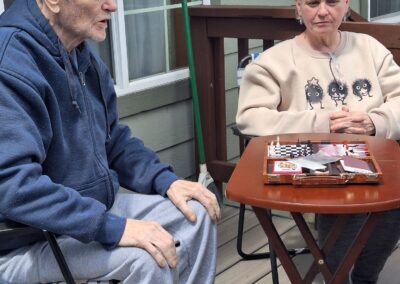 Two people sit on a patio, one wearing a blue hoodie and grey pants, the other in a beige sweater with a graphic design. They are at a small table with magazines and paper.