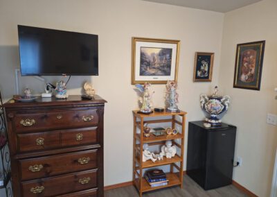 A room corner with a wooden dresser, a wall-mounted TV, framed art, figurines, a small bookshelf with decorations, and a mini-fridge.